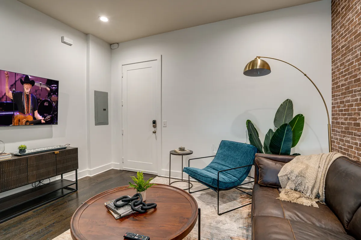 A modern living room featuring a brown leather sofa and blue accent chair, arranged around a wooden coffee table. A large television displays a live performance, and lush indoor plants add warmth. The setting is comfortable with ample space for group gatherings, movie nights, or relaxed conversation.