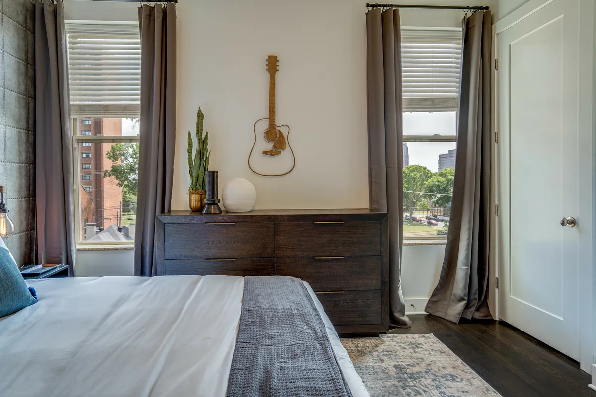 This image captures a king bedroom featuring a bed with blue accents, a green plant, a dark wooden dresser, and a guitar mounted on the wall. The space is bathed in soft, natural light, creating a tranquil and creative atmosphere. The furnishings and decor reflect modern elegance and comfort, aligning with the section's emphasis on restorative sleep and peaceful downtime.