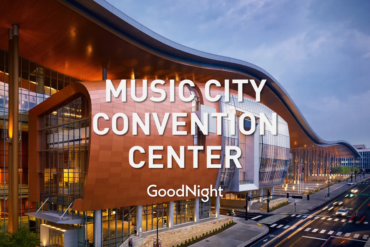 A contemporary exterior of the Music City Center, with its striking curved architecture, floor-to-ceiling windows, and illuminated signage shining in the evening. The area appears lively and sophisticated, hinting at ongoing events and city activity.