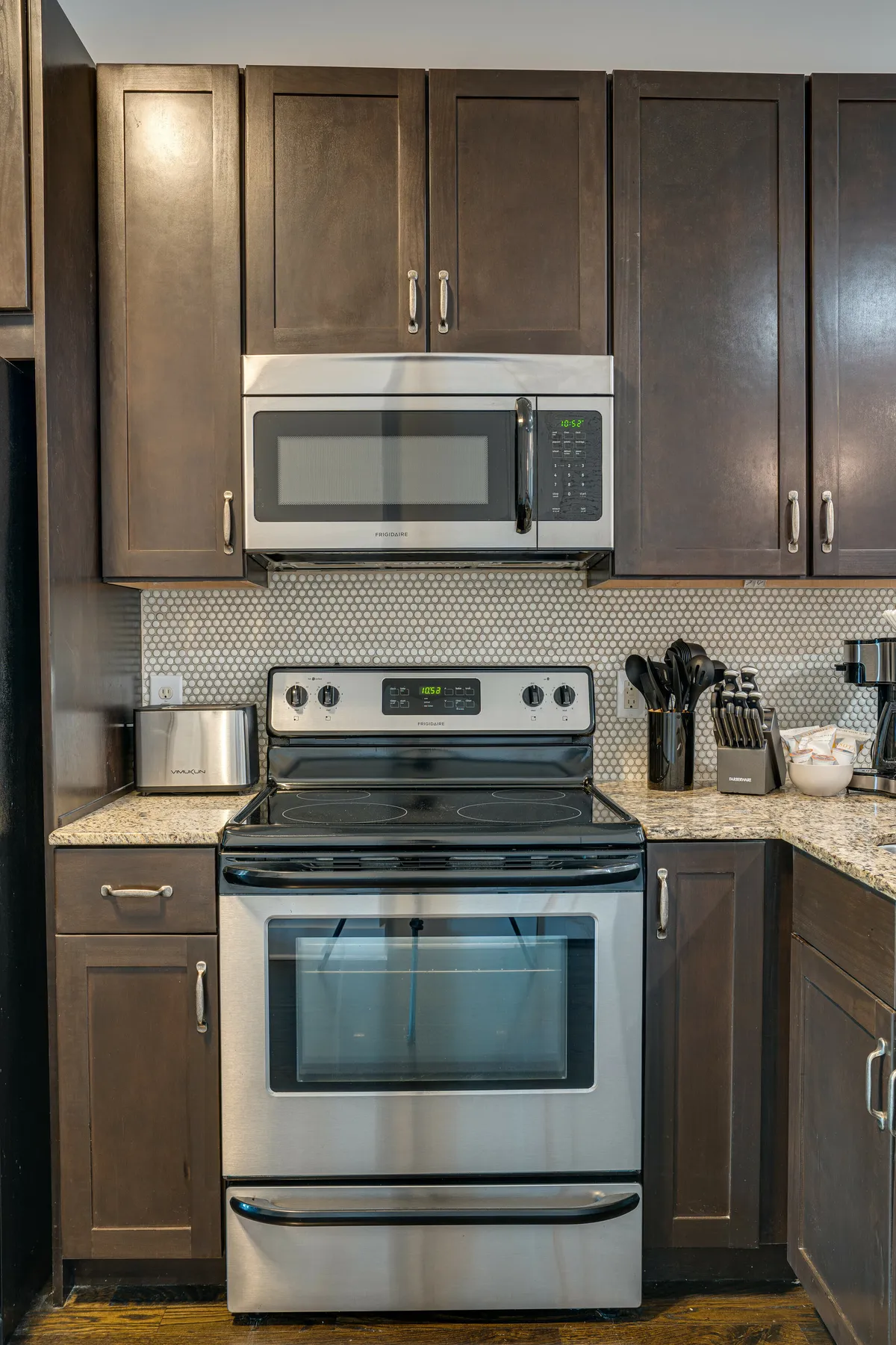 A cozy, functional kitchen featuring stainless steel appliances, a granite countertop, a modern microwave, and a kitchen utensils holder. The space is organized and ready for meal preparation, with visible cookware and tools that suggest it's equipped for everything from quick snacks to elaborate dinners.