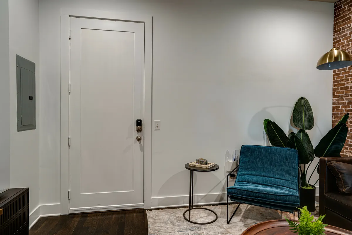 This image captures the entryway of the home, featuring a welcoming doorway, a blue accent chair, a small side table, a potted plant, and a backdrop of exposed brick wall. The scene feels cozy and inviting, setting a positive first impression for arriving guests.