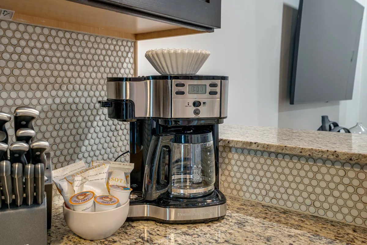 A close-up of a coffee maker, a set of knives, and a bowl with coffee pods on a kitchen countertop. The scene exudes morning comfort, suggesting the coffee maker is ready for use and breakfast is moments away.