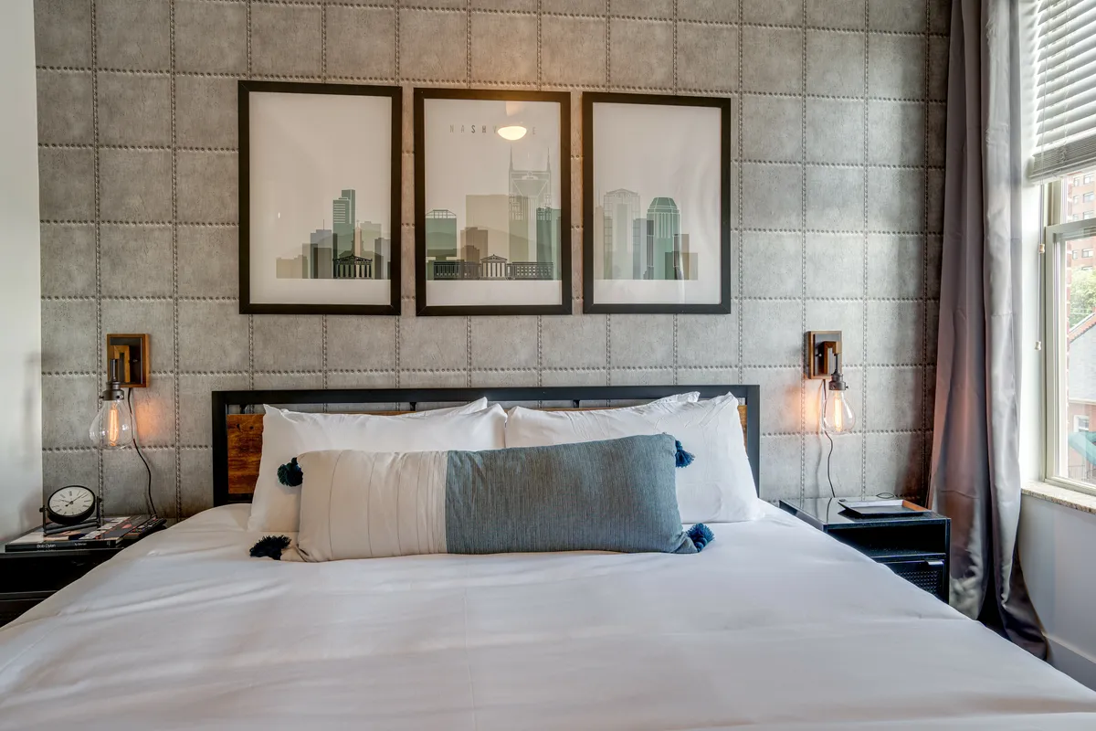 A stylish bedroom scene with a king bed adorned with decorative pillows, framed wall art above the headboard, and soft bedside lighting. The space feels inviting and serene, ideal for relaxation and gentle mornings.