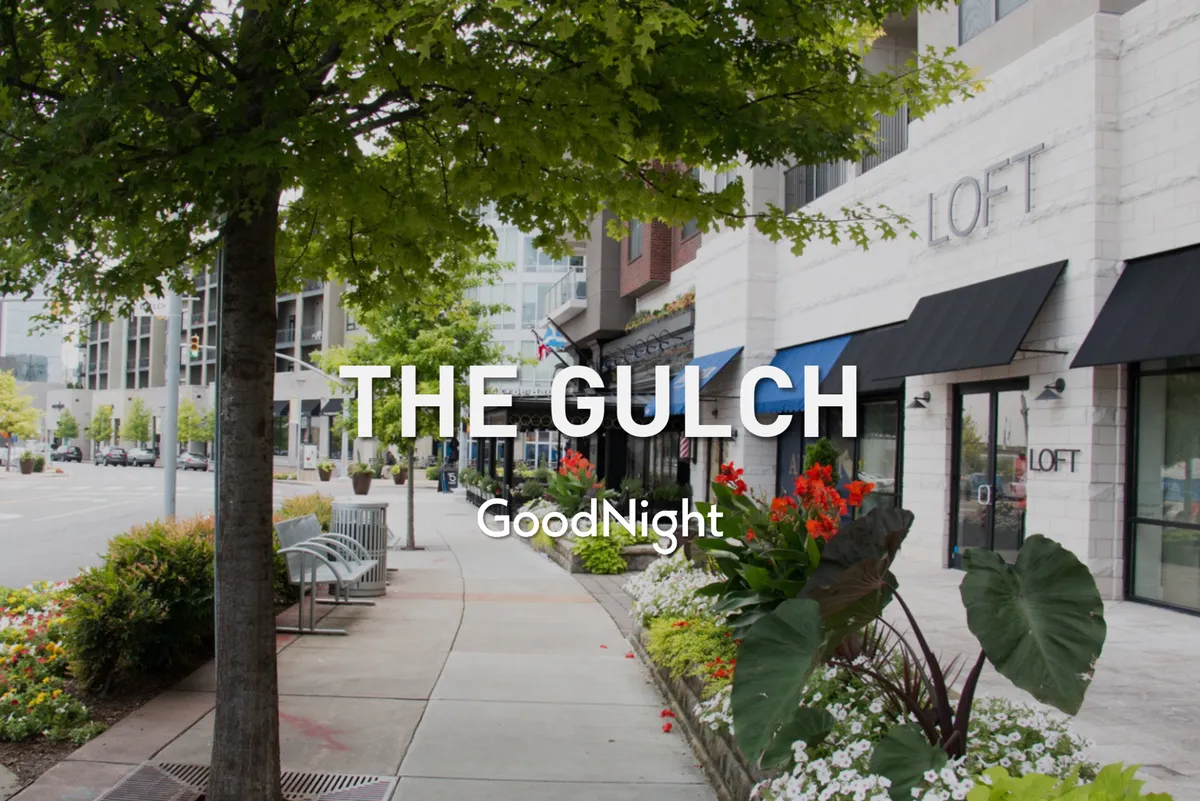 A lively, tree-lined sidewalk with vibrant storefronts, flower beds, and a welcoming seating area. The image captures a safe, family-friendly urban neighborhood ideal for strolls with children. The abundance of greenery offers a sense of calm and casual community energy.