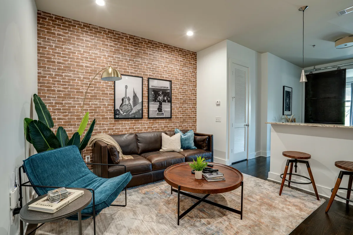 The image showcases a chic, open-concept living area with a striking exposed brick wall, plush brown leather sofa, stylish blue accent chair, modern round coffee table, lush green plant, and artistic black-and-white photographs. The scene radiates warmth and invites guests to relax, socialize, or unwind in comfort.